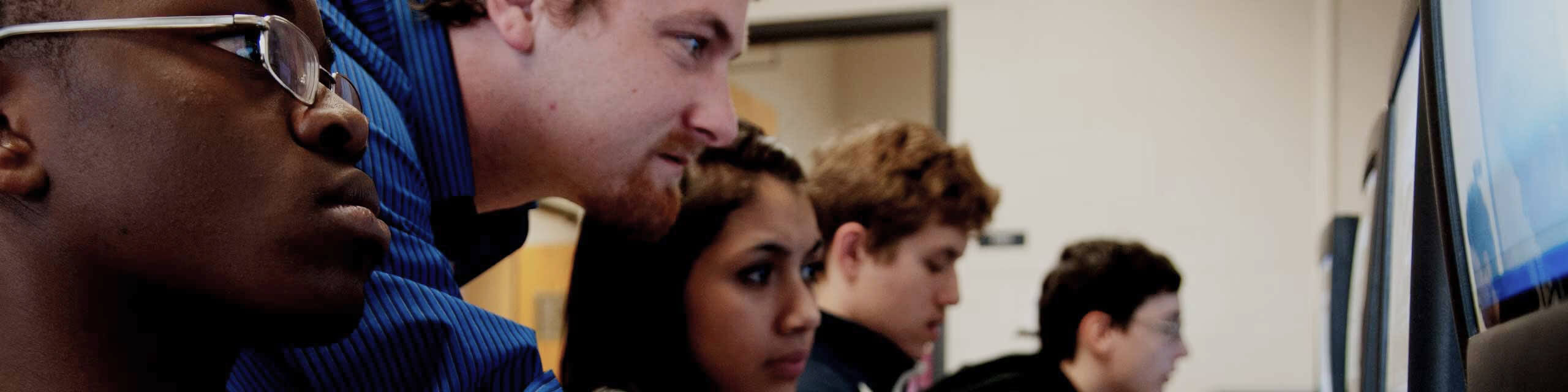 Students and teacher looking to a computer screen on a computer lab room.